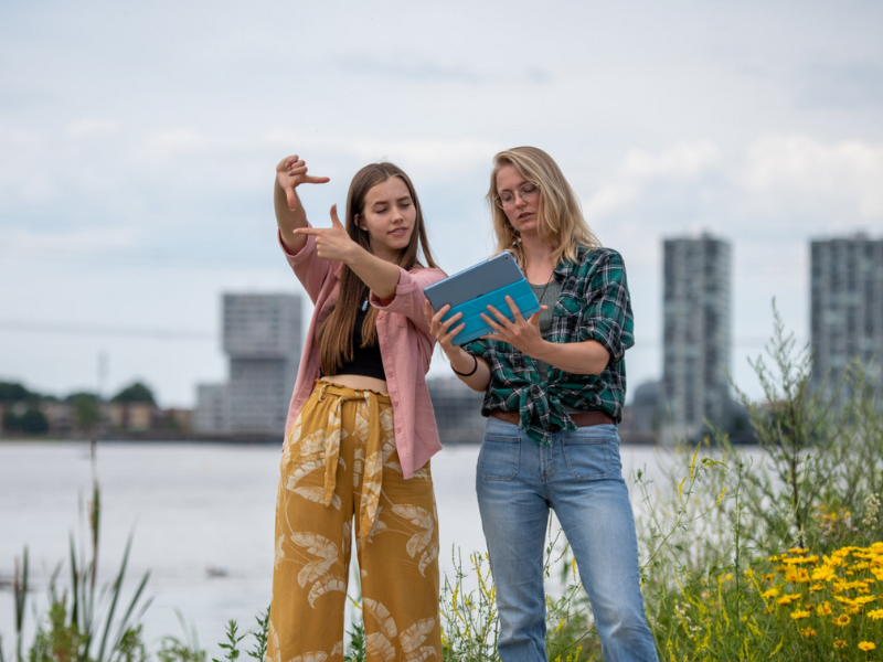 Twee meiden buiten bij het water in Almere met gebouwen op de achtergrond houden een kaart vast en praten over landschapsinrichting