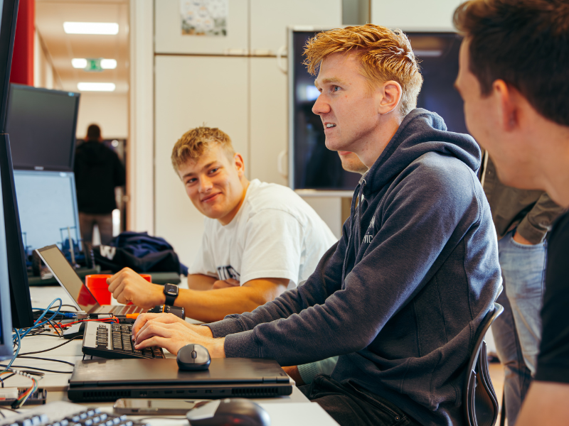 Techniek studenten achter laptops. 3 jongens. 1 kijkt naar het scherm, 1 kijkt naar de ander en lacht